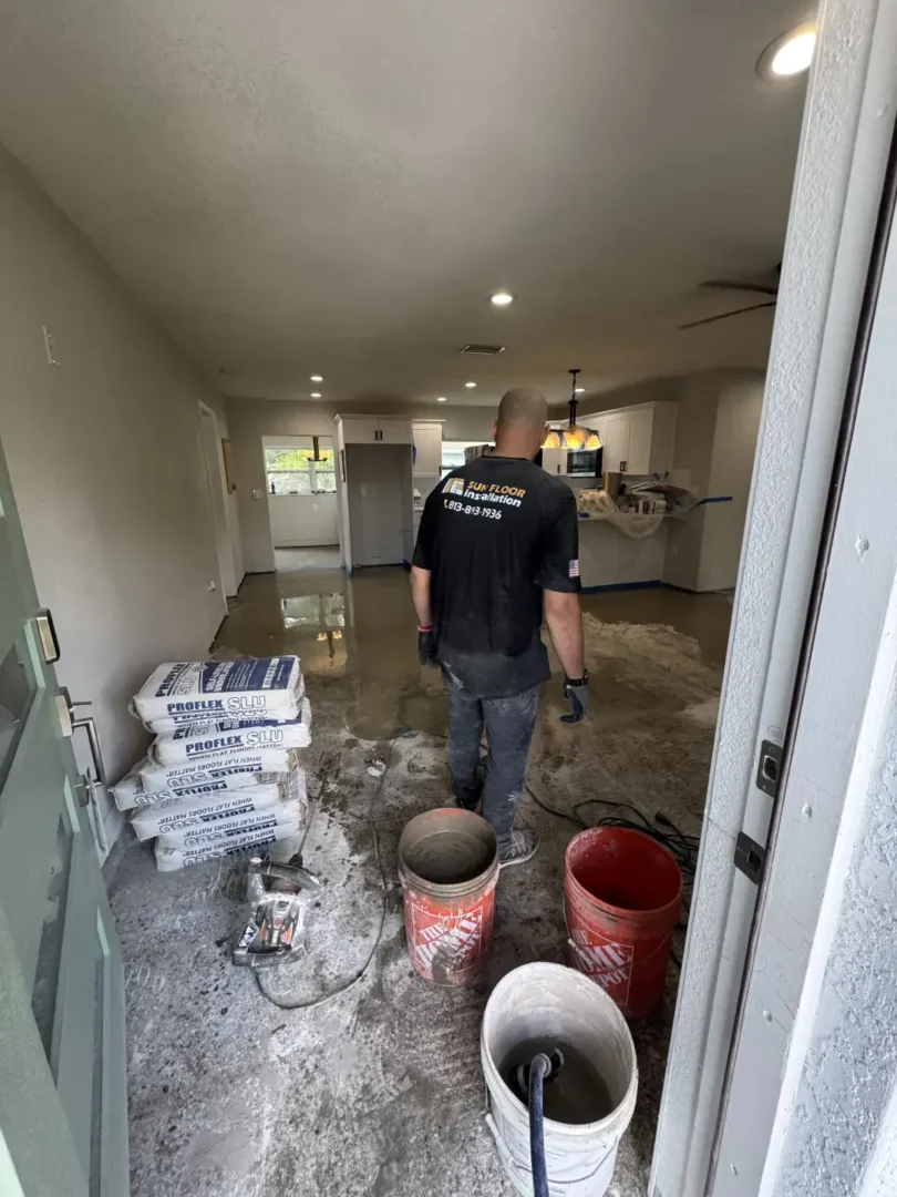 Worker leveling concrete floor inside a residential home with flooring materials and tools near garage entrance