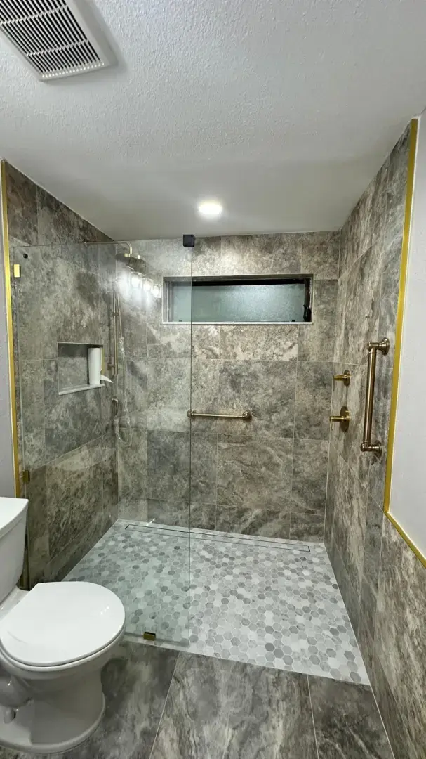 Let's Transform Your Bathroom Together Walk-in shower with a glass door, gray stone tile, brass grab bars, and hexagon mosaic floor in a bathroom